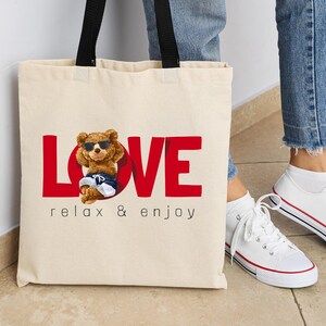 May include: A cream tote bag with black handles. The bag displays the red word "LOVE" with a cartoon bear wearing sunglasses and shorts. Underneath the word "LOVE" is the text "relax & enjoy".