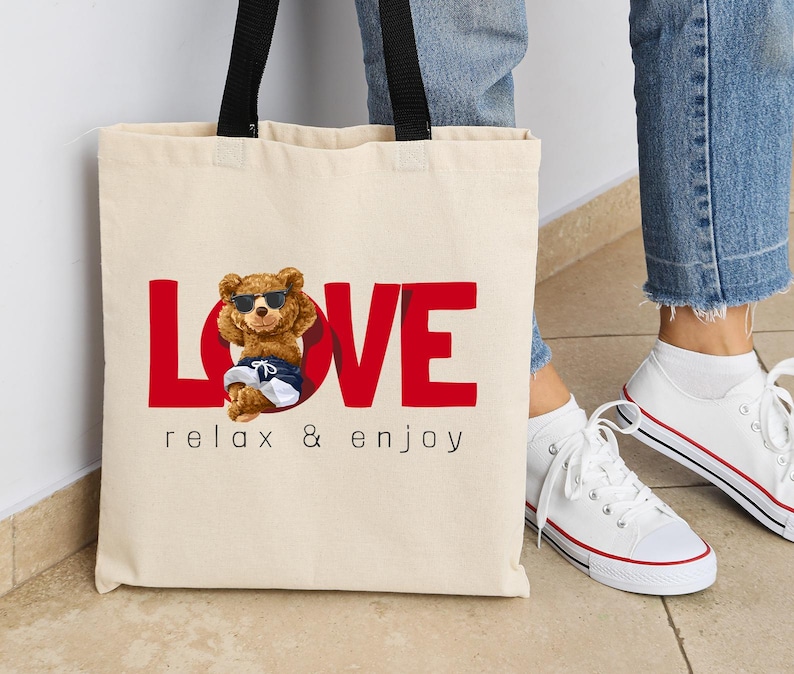 May include: A cream tote bag with black handles. The bag displays the red word "LOVE" with a cartoon bear wearing sunglasses and shorts. Underneath the word "LOVE" is the text "relax & enjoy".