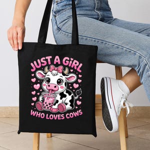 May include: Black tote bag with a cartoon cow design, drinking from a cup. The text "JUST A GIRL WHO LOVES COWS" is printed in pink. The bag has black handles. The cow has a pink bow.