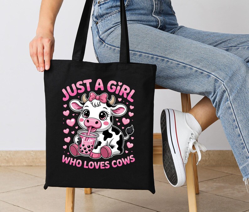 May include: Black tote bag with a cartoon cow design, drinking from a cup. The text "JUST A GIRL WHO LOVES COWS" is printed in pink. The bag has black handles. The cow has a pink bow.