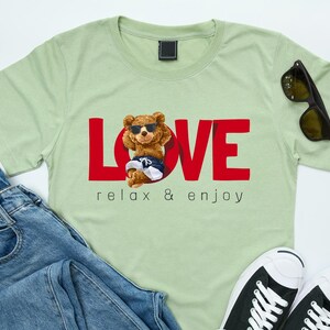 May include: Pale green t-shirt with the word "LOVE" in red, featuring a cartoon bear wearing sunglasses and shorts. The text "relax & enjoy" is below the bear. Sunglasses and blue jeans are next to the shirt.