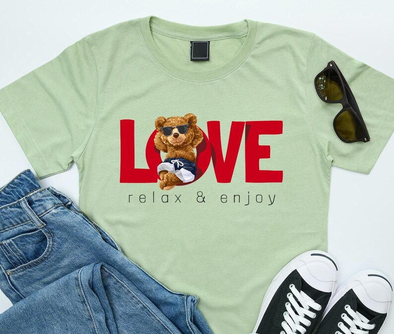 May include: Pale green t-shirt with the word "LOVE" in red, featuring a cartoon bear wearing sunglasses and shorts. The text "relax & enjoy" is below the bear. Sunglasses and blue jeans are next to the shirt.