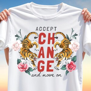 Puede incluir: Camiseta blanca con un diseño que presenta dos tigres, rosas y las palabras "ACCEPT CHANGE and move on" en rojo y negro. Los tigres son naranjas y negros, y las rosas son rosas.