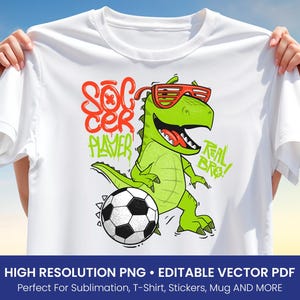 Op de afbeelding: Wit T-shirt met een cartoon dinosaurus met een zonnebril en een voetbal. Het ontwerp bevat de woorden "Soccer Player" in oranje en groen, met "Real Bro!" in groen.