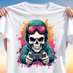 May include: White t-shirt featuring a skull wearing a teal hoodie and red-tinted goggles, holding a video game controller. The design includes colorful paint splatters in shades of orange, pink, and purple. The shirt is held up by hands.