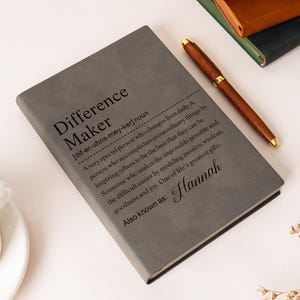 May include: Gray journal with the text "Difference Maker" and a definition. A wooden pen with gold accents rests on the journal. A cup of coffee is in the lower left corner. The journal is personalized with the name "Hannah".