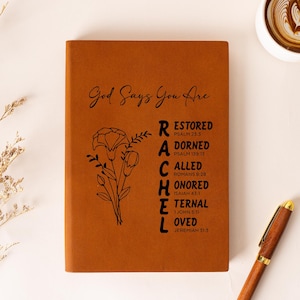 May include: A brown leather journal with the words "God Says You Are" and "RACHEL" printed on the cover. The journal also features a floral illustration and the words "RESTORED, DORNED, CALLED, HONORED, ETERNAL, LOVED" with corresponding Bible verses. A pen and a cup of coffee are also in the image.
