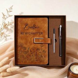 May include: A tan leather-effect journal with a gold-toned metal clasp, featuring the text "Bella's NEW CHAPTER" and a black line-art floral design. The journal is presented in a brown box alongside a black pen and a silver-toned pen refill.