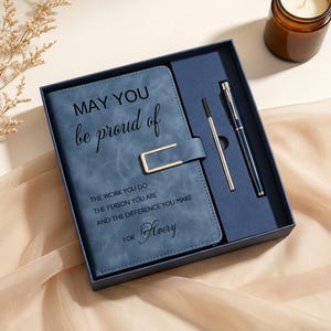 Op de afbeelding: Een blauw notitieboek met een goudkleurige metalen sluiting en een bijpassende pen in een donkerblauwe geschenkdoos. De kaft bevat de tekst: MAY YOU be proud of THE WORK YOU DO THE PERSON YOU ARE AND THE DIFFERENCE YOU MAKE FOR Avery in zwart.