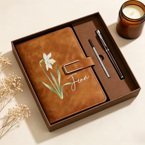 May include: A brown notebook with a white daffodil illustration and the name Jean printed in white script, featuring a gold-tone metal clasp. The notebook is presented in a brown gift box alongside a matching pen and a refill, near a candle.