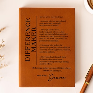 Könnte beinhalten: Ein rechteckiges Notizbuch mit einem rostorangen Einband und schwarzem Text, der das Substantiv "DIFFERENCE MAKER" in vier nummerierten Punkten definiert, ergänzt durch "see also: Denver" in Schreibschrift und einen Stift in Holzoptik.