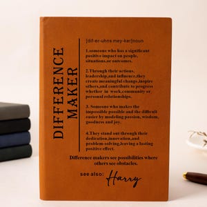May include: A rectangular, burnt-orange notebook featuring a dictionary-style definition of the term "DIFFERENCE MAKER" printed in black text, with the name "Harry" written in a cursive script at the bottom under the "see also" section.