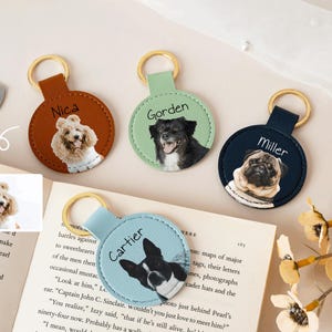 May include: A collection of personalized pet keychains. Each keychain features a circular leather tag with a gold-colored ring. The tags display custom portraits of dogs, with names like "Nica," "Gorden," "Miller," and "Cartier" printed above the images.