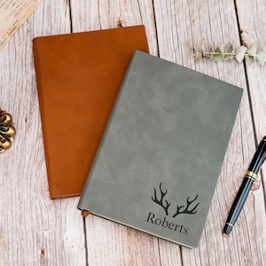 Puede incluir: Dos cuadernos de cuero, uno marrón y otro gris, descansan sobre una superficie de madera. El cuaderno gris presenta un diseño de cuernos de ciervo y el nombre "Roberts" en negro. Un bolígrafo negro con detalles dorados está cerca.