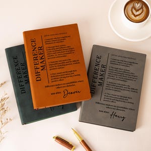 K&ouml;nnte beinhalten: Drei rechteckige Notizb&uuml;cher in Dunkelgr&uuml;n, Hellbraun und Grau, die jeweils eine schwarz gedruckte Definition des Begriffs Difference Maker aufweisen. Das hellbraune Notizbuch enth&auml;lt den Namen Denver und das graue den Namen Harry in Schreibschrift.