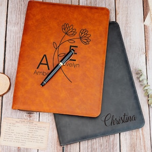 Puede incluir: Se muestran dos diarios encuadernados en cuero, uno en marrón cálido y otro en gris oscuro. El diario marrón presenta un diseño floral y los nombres "Amber" y "Evelyn". Un bolígrafo negro descansa encima. El diario gris está inscrito con el nombre "Christina".