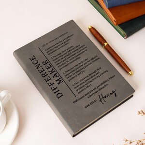K&ouml;nnte beinhalten: Ein graues Notizbuch mit der Definition des Wortes DIFFERENCE MAKER in schwarzem Druck, inklusive nummerierter Punkte und dem Namen Harry in Schreibschrift am unteren Rand. Ein brauner Stift liegt diagonal auf dem Einband.