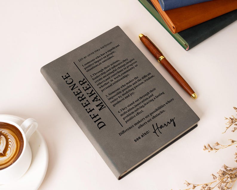 K&ouml;nnte beinhalten: Ein graues Notizbuch mit der Definition des Wortes DIFFERENCE MAKER in schwarzem Druck, inklusive nummerierter Punkte und dem Namen Harry in Schreibschrift am unteren Rand. Ein brauner Stift liegt diagonal auf dem Einband.