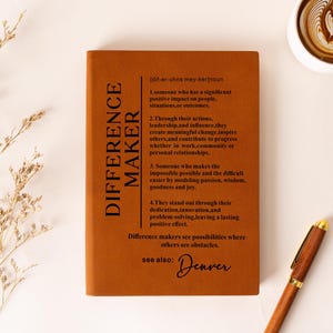 K&ouml;nnte beinhalten: Ein rechteckiges Notizbuch mit einem rostorangen Einband und schwarzem Text, der das Substantiv "DIFFERENCE MAKER" in vier nummerierten Punkten definiert, erg&auml;nzt durch "see also: Denver" in Schreibschrift und einen Stift in Holzoptik.