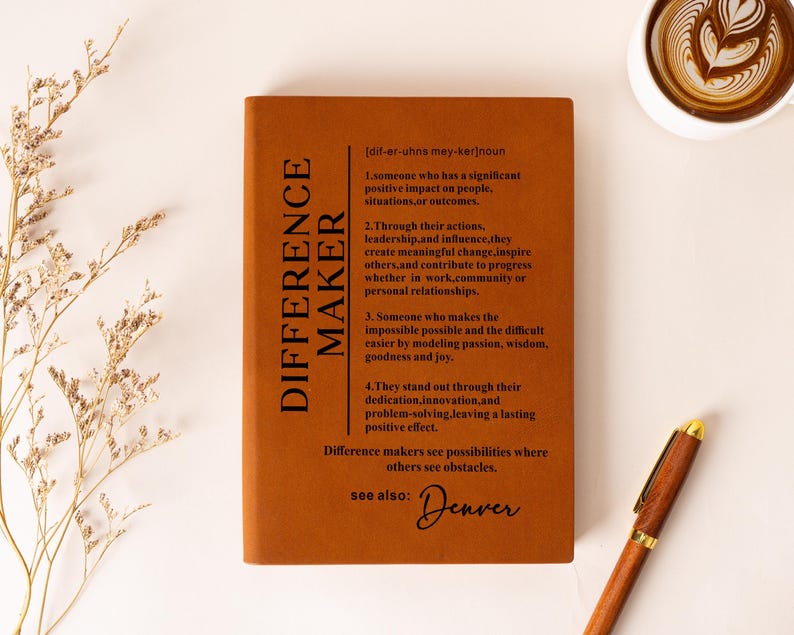K&ouml;nnte beinhalten: Ein rechteckiges Notizbuch mit einem rostorangen Einband und schwarzem Text, der das Substantiv "DIFFERENCE MAKER" in vier nummerierten Punkten definiert, erg&auml;nzt durch "see also: Denver" in Schreibschrift und einen Stift in Holzoptik.