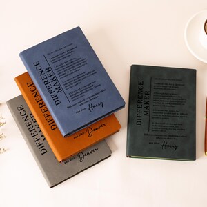 K&ouml;nnte beinhalten: Vier rechteckige Notizb&uuml;cher mit matten Einb&auml;nden in Blau, Orange, Grau und Dunkelgr&uuml;n. Jeder Einband zeigt den Text "DIFFERENCE MAKER" und eine Definition sowie einen personalisierten Namen. Ein brauner Stift und eine Tasse Kaffee liegen daneben.