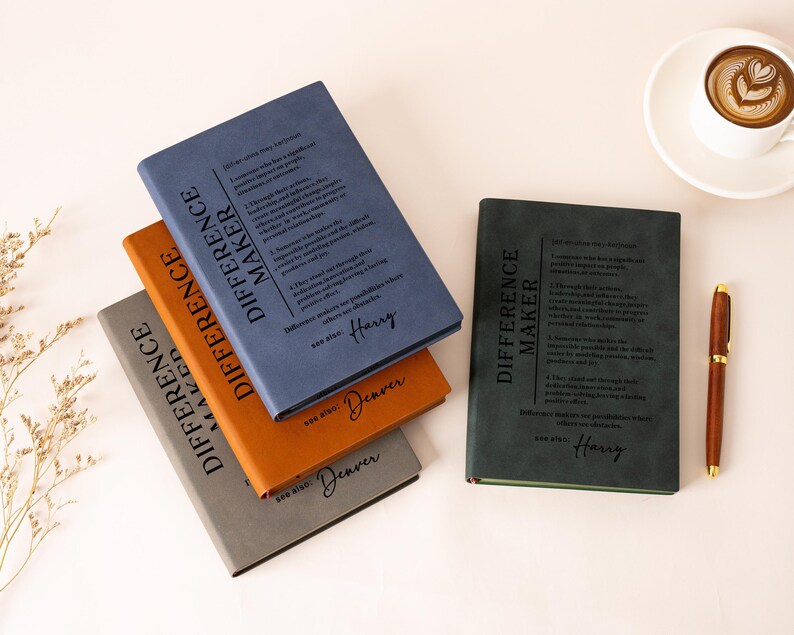 K&ouml;nnte beinhalten: Vier rechteckige Notizb&uuml;cher mit matten Einb&auml;nden in Blau, Orange, Grau und Dunkelgr&uuml;n. Jeder Einband zeigt den Text "DIFFERENCE MAKER" und eine Definition sowie einen personalisierten Namen. Ein brauner Stift und eine Tasse Kaffee liegen daneben.