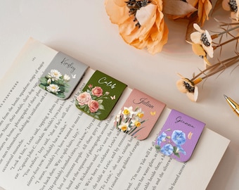 May include: Four personalized leather bookmarks with floral designs and names: Kingsley, Caleb, Julian, and Gianna. Each bookmark features a different flower: water lilies, roses, daffodils, and morning glories. The bookmarks are on a book page.