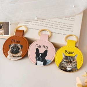 Puede incluir: Tres llaveros personalizados para mascotas. Los llaveros son redondos y tienen un aspecto similar al cuero. Cada llavero presenta un retrato de mascota diferente: un pug, un bulldog francés y un gato atigrado. Los llaveros tienen anillos de metal dorados y los nombres de las mascotas.