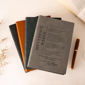 K&ouml;nnte beinhalten: Ein Stapel aus vier rechteckigen Notizb&uuml;chern mit matten Einb&auml;nden in Grau, Dunkelgr&uuml;n, Orange und Schwarz. Das oberste graue Notizbuch zeigt eine w&ouml;rterbuchartige Definition des Begriffs Difference Maker in schwarzem Text, endend mit Watson.