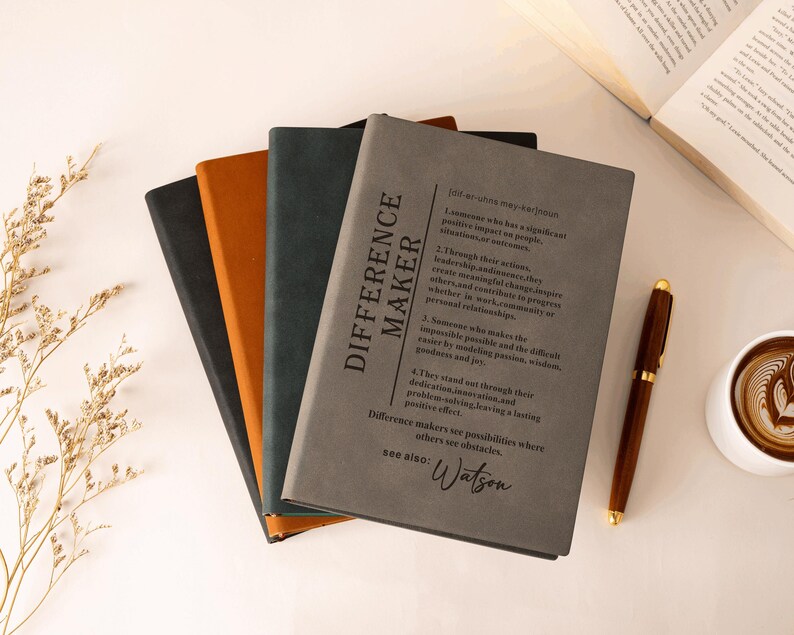 K&ouml;nnte beinhalten: Ein Stapel aus vier rechteckigen Notizb&uuml;chern mit matten Einb&auml;nden in Grau, Dunkelgr&uuml;n, Orange und Schwarz. Das oberste graue Notizbuch zeigt eine w&ouml;rterbuchartige Definition des Begriffs Difference Maker in schwarzem Text, endend mit Watson.