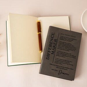 K&ouml;nnte beinhalten: Ein offenes Notizbuch mit cremefarbenen Seiten und einem dunkelbraunen Holzstift darin. Daneben liegt ein geschlossenes dunkelgraues Notizbuch mit dem aufgedruckten Text 'DIFFERENCE MAKER', gefolgt von einer w&ouml;rterbuchartigen Definition und dem Wort 'Denver'.