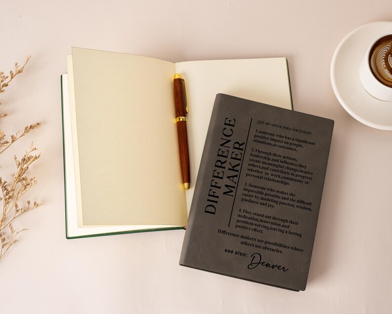 K&ouml;nnte beinhalten: Ein offenes Notizbuch mit cremefarbenen Seiten und einem dunkelbraunen Holzstift darin. Daneben liegt ein geschlossenes dunkelgraues Notizbuch mit dem aufgedruckten Text 'DIFFERENCE MAKER', gefolgt von einer w&ouml;rterbuchartigen Definition und dem Wort 'Denver'.