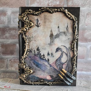 Könnte beinhalten: Ein Fantasy-Tagebuch mit einer Drachenillustration. Das Cover zeigt ein gerahmtes Bild eines Drachen, einer Burg und einer Landschaft mit goldenen Verzierungen. Das Tagebuch ist dunkel mit einem Verschluss.
