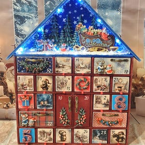 Puede incluir: Un calendario de Adviento navide&ntilde;o de madera con forma de casa y veinticuatro cajones numerados. El techo azul incluye luces LED, pinos y un trineo de Santa. Cada caj&oacute;n muestra ilustraciones festivas con ratones, regalos y escenas invernales, junto con tiradores met&aacute;licos decorativos.