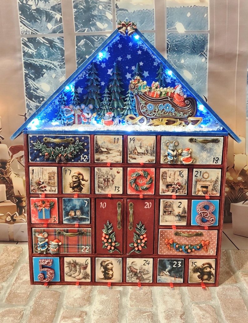 Puede incluir: Un calendario de Adviento navide&ntilde;o de madera con forma de casa y veinticuatro cajones numerados. El techo azul incluye luces LED, pinos y un trineo de Santa. Cada caj&oacute;n muestra ilustraciones festivas con ratones, regalos y escenas invernales, junto con tiradores met&aacute;licos decorativos.