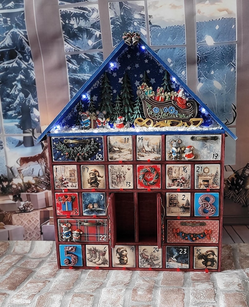 Puede incluir: Un calendario de Adviento de madera con forma de casa y veinticuatro cajones numerados. El techo presenta un fondo azul estrellado, pinos en miniatura y un trineo de Santa. Cada caj&oacute;n muestra ilustraciones festivas, incluyendo ratones, regalos y escenas navide&ntilde;as, con algunos cajones que presentan detalles decorativos tridimensionales.