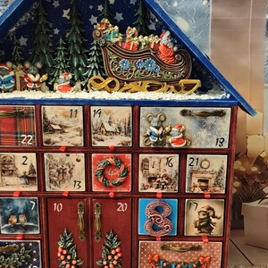 Puede incluir: Un calendario de adviento de madera con forma de casa y techo azul, que cuenta con veinticuatro cajones numerados decorados con ilustraciones festivas de ratones, Santa Claus y escenas invernales. La parte superior muestra un trineo en relieve.