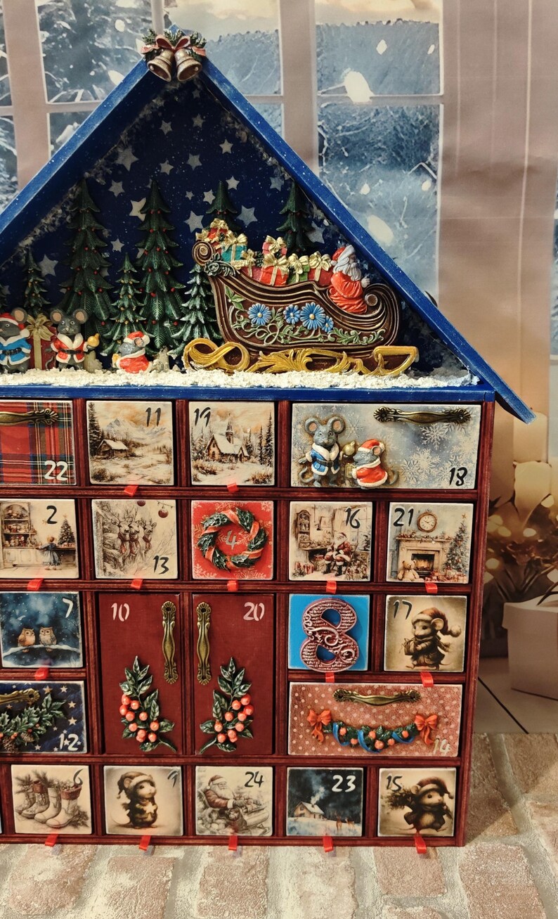Puede incluir: Un calendario de adviento de madera con forma de casa y techo azul, que cuenta con veinticuatro cajones numerados decorados con ilustraciones festivas de ratones, Santa Claus y escenas invernales. La parte superior muestra un trineo en relieve.