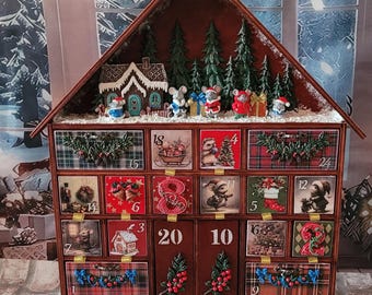 May include: A wooden house-shaped advent calendar with twenty-four numbered drawers. The top features a miniature scene with green pine trees, a small house, and several mouse figurines. Each drawer displays unique holiday-themed illustrations.