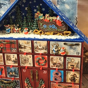 Puede incluir: Un calendario de adviento navide&ntilde;o de madera con forma de casa y techo azul, que cuenta con veinticuatro cajones numerados decorados con escenas festivas, ratones y motivos navide&ntilde;os, rematado con un trineo en miniatura, &aacute;rboles y campanas doradas.