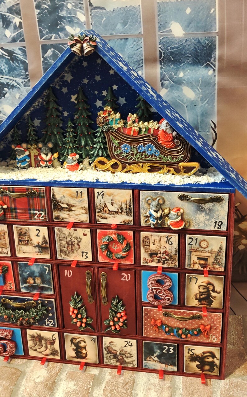 Puede incluir: Un calendario de adviento navide&ntilde;o de madera con forma de casa y techo azul, que cuenta con veinticuatro cajones numerados decorados con escenas festivas, ratones y motivos navide&ntilde;os, rematado con un trineo en miniatura, &aacute;rboles y campanas doradas.