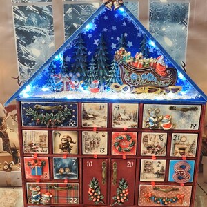 Puede incluir: Un calendario de adviento de madera con forma de casa, veinticuatro cajones numerados y dos puertas centrales. El techo azul tiene luces LED, un bosque en miniatura y un trineo. Cada frente de caj&oacute;n muestra im&aacute;genes festivas y adornos.