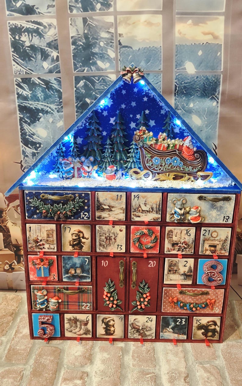 Puede incluir: Un calendario de adviento de madera con forma de casa, veinticuatro cajones numerados y dos puertas centrales. El techo azul tiene luces LED, un bosque en miniatura y un trineo. Cada frente de caj&oacute;n muestra im&aacute;genes festivas y adornos.