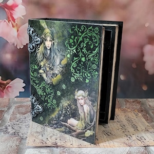 Puede incluir: Una caja rectangular de madera negra diseñada para parecer un libro, con una cubierta verde oscuro que presenta ilustraciones de dos figuras élficas, intrincados patrones de enredaderas verdes y detalles metálicos plateados en las esquinas.