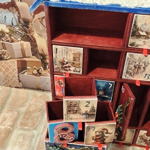 Puede incluir: Un calendario de adviento de madera con techo azul y compartimentos interiores rojos. El frente presenta cajones numerados decorados con ilustraciones festivas, incluyendo a Santa, ratones y escenas invernales. Peque&ntilde;as figuras de ratones con ropa festiva.