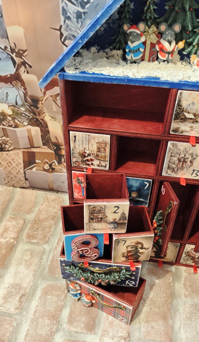 Puede incluir: Un calendario de adviento de madera con techo azul y compartimentos interiores rojos. El frente presenta cajones numerados decorados con ilustraciones festivas, incluyendo a Santa, ratones y escenas invernales. Peque&ntilde;as figuras de ratones con ropa festiva.