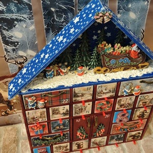 Puede incluir: Un calendario de adviento de madera con forma de casa y techo azul con estrellas blancas. La parte superior muestra una escena en miniatura con pinos verdes, un trineo y ratones. El frente tiene veinticuatro cajones numerados con ilustraciones.