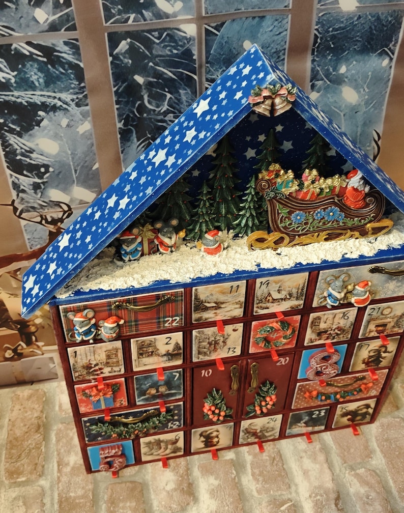 Puede incluir: Un calendario de adviento de madera con forma de casa y techo azul con estrellas blancas. La parte superior muestra una escena en miniatura con pinos verdes, un trineo y ratones. El frente tiene veinticuatro cajones numerados con ilustraciones.