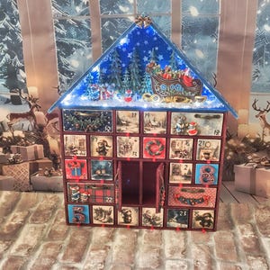 Puede incluir: Un calendario de adviento de madera en forma de casa con veinticuatro cajones numerados, con un techo a dos aguas con un cielo azul estrellado, &aacute;rboles de hoja perenne y una escena de trineo de Santa. Los cajones muestran ilustraciones navide&ntilde;as.
