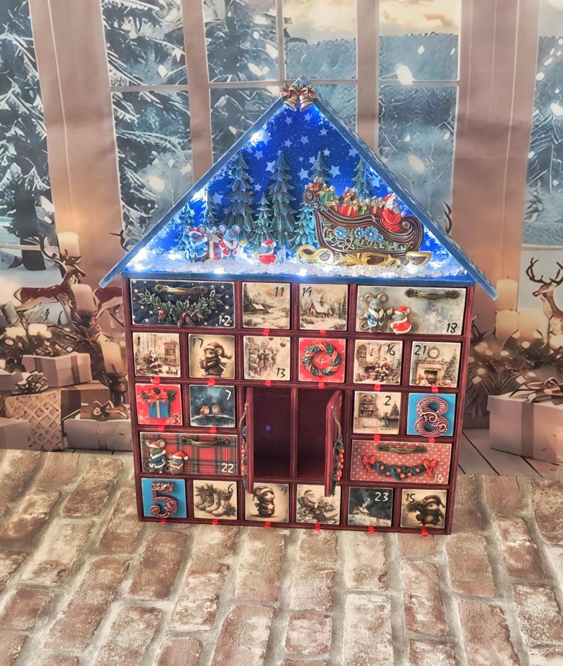 Puede incluir: Un calendario de adviento de madera en forma de casa con veinticuatro cajones numerados, con un techo a dos aguas con un cielo azul estrellado, &aacute;rboles de hoja perenne y una escena de trineo de Santa. Los cajones muestran ilustraciones navide&ntilde;as.